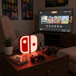 Op de afbeelding: Een rood-witte verlichte gaming controller lamp in de vorm van een joy-con, geplaatst op een houten tafel naast een zwarte draadloze gamecontroller, een zwarte koptelefoon, een glas water en een videospelhoesje.