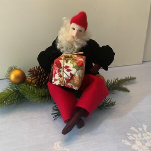 May include: A seated Christmas figurine with a red hat and pants, black jacket, and white beard, holding a wrapped gift. The figurine is surrounded by pine needles, a gold ornament, and a pine cone.