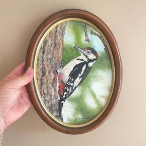 Peut inclure: Une peinture ovale représentant un pic perché sur un tronc d'arbre texturé. L'oiseau présente un plumage noir, blanc et rouge avec un bec pointu. L'arrière-plan montre un feuillage vert doux. Le cadre en bois a une finition brun foncé et dorée.