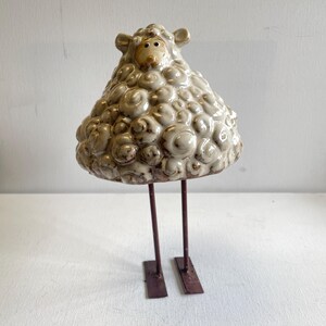 May include: A ceramic sheep figurine with a textured, off-white body and a light brown face. The sheep stands on two thin, dark brown metal legs attached to a rectangular base. The sheep's woolly body is detailed with rounded, textured patterns.