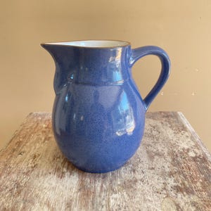 Può includere: Una brocca in ceramica con smalto blu maculato e un corpo liscio e arrotondato. Il recipiente presenta un ampio beccuccio, un manico curvo sul lato destro e una finitura interna bianca. La superficie esterna ha un tono blu uniforme.