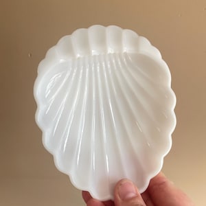 Op de afbeelding: Een witte schaal van melkglas in de vorm van een sint-jakobsschelp. De schaal heeft een gladde, glanzende afwerking met verhoogde, stralende ribbels die van de basis naar de geschulpte randen lopen, wat een schelpachtige vorm creëert.