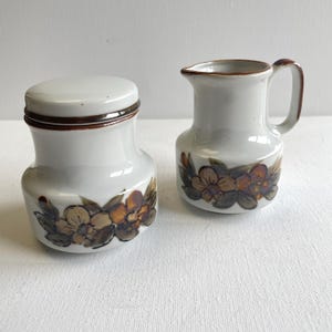 Op de afbeelding: Een witte keramische set van een melkkan en een pot met deksel. Beide stukken hebben een bruin bloemmotief met oranje en bruine bloemen en groene bladeren. De randen en het handvat van de melkkan zijn bruin.