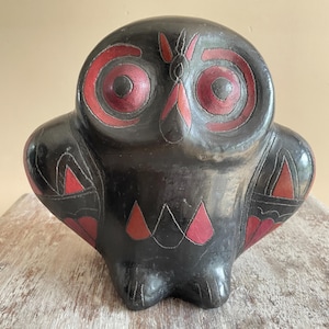 Puede incluir: Una figura de búho de cerámica negra con prominentes ojos circulares rojos y patrones geométricos rojos en las alas y el pecho. La superficie tiene un acabado oscuro y liso con líneas grabadas que delinean los elementos decorativos.