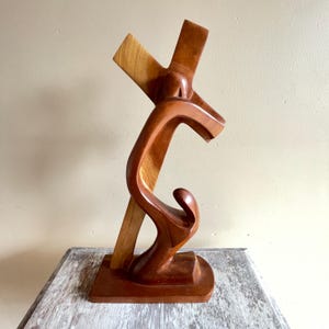 Peut inclure: Sculpture en bois représentant une figure portant une croix. La croix est en bois clair, contrastant avec le bois brun-rouge plus foncé de la figure. La sculpture a une base carrée et est un objet décoratif.