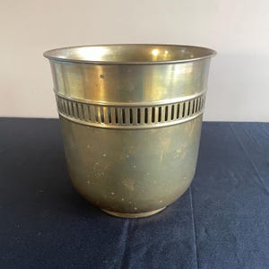 Peut inclure: Un pot en laiton vintage de forme cylindrique, orné d'une bande décorative à fentes verticales. Le pot présente une finition légèrement ternie, lui conférant un aspect classique et vieilli. L'intérieur est lisse et le bord est arrondi.