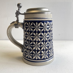 Peut inclure: Une chope à bière en céramique avec un corps bleu profond orné d'un motif géométrique en étoile blanc en relief. Le récipient comprend une anse gris clair et un couvercle métallique articulé assorti avec un bouton sphérique au sommet.