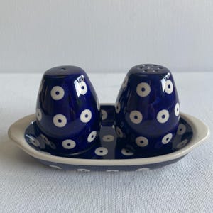 Peut inclure: Un ensemble salière et poivrière en céramique avec un glaçage bleu foncé et des pois blancs. L'ensemble comprend deux salières et un plateau ovale assorti avec un bord crème. Les salières ont une forme arrondie.