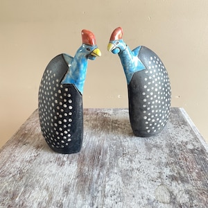 Può includere: Due statuette di faraone in ceramica con corpo nero a pois bianchi, collo blu brillante, creste rosse e becchi gialli. Gli uccelli sono posizionati l'uno di fronte all'altro, mostrando una forma liscia e arrotondata con dettagli dipinti.