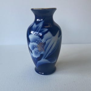 Könnte beinhalten: Kleine, dunkelblaue Keramikvase mit Goldrand. Die Vase hat ein weißes und goldenes Blumendesign. Die Vase ist etwa 15 cm hoch und hat einen abgerundeten Körper.