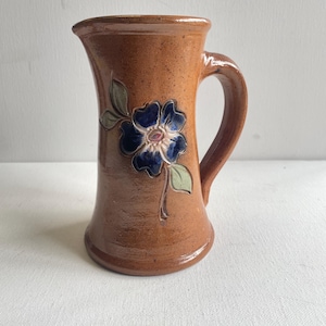 Peut inclure: Une cruche en céramique brune avec une finition brillante et une texture tachetée. La cruche présente un motif de fleur bleue en relief peint à la main avec des feuilles vertes sur le devant, un bord large et une anse incurvée sur le côté.