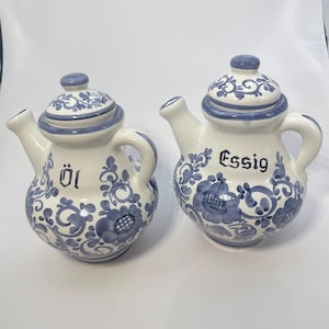 Peut inclure: Deux huiliers-vinaigriers en céramique blanche avec des motifs floraux bleus et des couvercles. Le cruet de gauche porte le mot "Öl" et celui de droite le mot "Essig" en lettres noires.