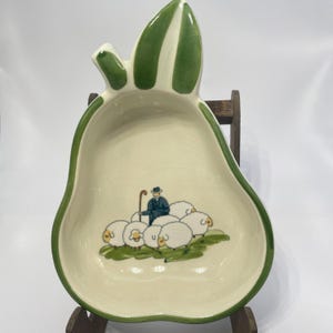 Puede incluir: Una fuente de cerámica en forma de pera con borde verde y parte superior en forma de hoja. El interior presenta una escena pastoral de un pastor y ovejas. La fuente es de color blanquecino con un acabado brillante.