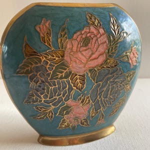 Op de afbeelding: Een turquoise vaas met een gouden rand en voet. De vaas heeft een bloemenontwerp met roze rozen, groene bladeren en gouden accenten. De vaas heeft een afgeronde vorm en een glad oppervlak.