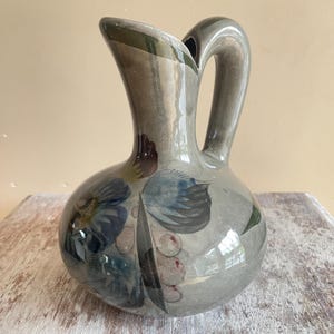 Puede incluir: Una jarra de cerámica gris brillante con cuerpo redondeado y asa curva. La superficie presenta un diseño floral pintado a mano en tonos azul, verde oscuro y rosa suave, con pinceladas visibles y un acabado liso y reflectante en toda la pieza.
