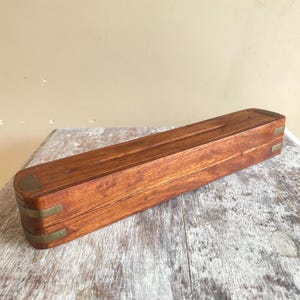 May include: A rectangular wooden box with a warm brown finish and visible wood grain. The box features rounded edges reinforced with polished brass corner accents and brass bands along the sides, providing a structured and durable appearance.