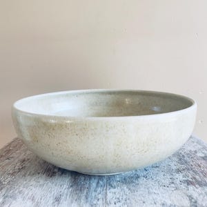 Könnte beinhalten: Eine breite, flache Keramikschale mit einem glatten, abgerundeten Rand. Die Außen- und Innenseite weisen eine gesprenkelte, cremeweiße Glasur mit subtilen beigen und hellbraunen Flecken auf, die der Oberfläche ein strukturiertes Aussehen verleihen.