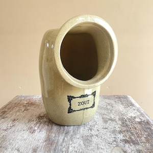 May include: A beige ceramic salt pig with a large opening and a curved design. The word "ZOUT" is printed in a black rectangular frame on the front. The salt pig is sitting on a weathered wooden surface.