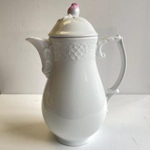 May include: A white porcelain coffee pot with a lid and handle. The lid is topped with a pink and white floral design. The pot has a curved spout and ornate detailing around the body.