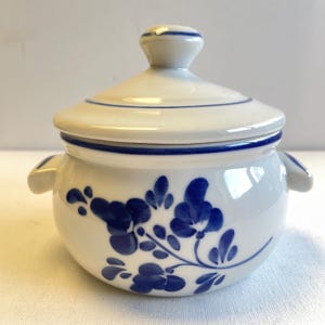 Peut inclure: Petit pot en céramique blanche avec couvercle et motif floral bleu. Le pot a deux petites poignées et un bouton sur le couvercle. Une bande bleue accentue le bord et le couvercle. Le motif floral est une fleur bleue stylisée.
