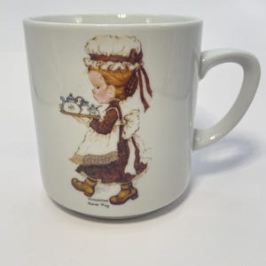 Pode incluir: Caneca de cerâmica branca com uma ilustração vintage de uma menina carregando uma bandeja com uma chaleira e xícaras. A menina está vestida com um chapéu e um avental. A caneca tem uma alça curva e o texto "Valentine Sarah Kay".