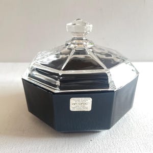 Peut inclure: Un pot en verre noir et transparent avec un couvercle à facettes. Le pot a une forme géométrique et une étiquette qui indique "arcocroc". Le couvercle est transparent et possède un bouton en forme de diamant.