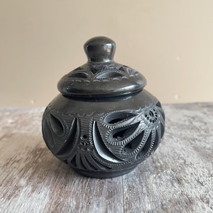 Può includere: Un barattolo in ceramica nera lucida di forma arrotondata con coperchio abbinato. La superficie presenta intricati intagli realizzati a mano e motivi floreali incisi che rivelano l'interno. Il contenitore ha una finitura liscia e brillante.