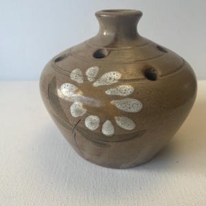 Peut inclure: Un vase en céramique marron avec un motif floral. Le vase a un corps arrondi avec un col étroit et plusieurs trous autour du dessus. Le motif floral est blanc avec des accents marron.
