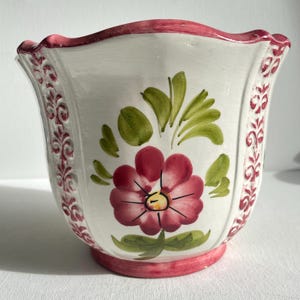 Pode incluir: Vaso de cerâmica branco com borda e base rosa onduladas. Apresenta um design floral pintado à mão, incluindo uma grande flor vermelha, folhas verdes e volutas decorativas vermelhas. Ideal para plantas pequenas.