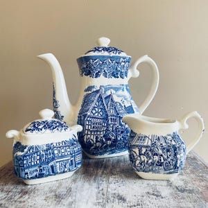 Pode incluir: Um conjunto de chá em cerâmica composto por um bule com tampa, um açucareiro com tampa e uma leiteira. Cada peça é de cor creme com um padrão azul detalhado que retrata cenas de aldeias históricas, carruagens puxadas por cavalos e bordas florais.