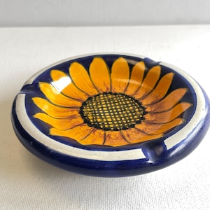 Może przedstawiać: Ceramiczna popielniczka z ręcznie malowanym wzorem słonecznika. Popielniczka ma w środku żywy żółty słonecznik, otoczony ciemnoniebieską obwódką z białymi akcentami. Popielniczka ma okrągły kształt z małym wycięciem na papierosy.