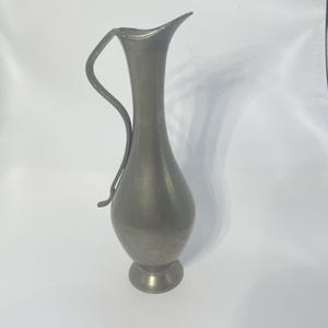 May include: A vintage pewter jug with a curved handle and a flared spout. The jug has a bulbous body and a round base. The metal has a dull, silver-grey finish. The jug is likely used for serving water or other beverages.