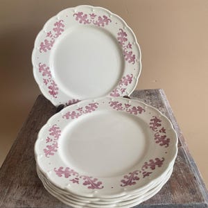 Op de afbeelding: Een stapel crèmekleurige keramische borden met een geschulpte rand en een herhalend mauvekleurig bloemmotief langs de rand. De borden hebben een gladde, glanzende afwerking en een consistent ontwerp over de gehele set serviesgoed.