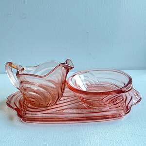 Peut inclure: Un ensemble de service à crème et sucrier en verre rose vintage. La crémière a une anse et un bec verseur, et le sucrier est rond. Les deux sont posés sur un plateau rectangulaire assorti avec un motif ondulé.