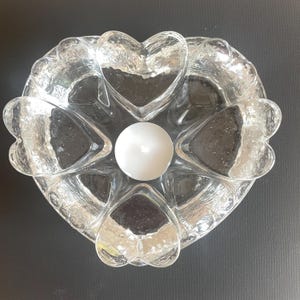 Puede incluir: Un portavelas de cristal transparente con forma de corazón grande, que presenta una superficie texturizada y ondulada. El diseño incorpora cinco hendiduras más pequeñas en forma de corazón dispuestas alrededor de un espacio circular central con una vela blanca.