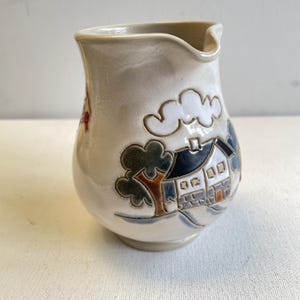 Peut inclure: Pichet en céramique crème avec un motif paysager. Le motif représente une maison blanche avec un toit bleu foncé, des arbres et des nuages. Le pichet a un bec courbé et un corps arrondi.