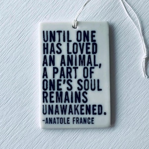 May include: A rectangular white ceramic tag with a quote in dark blue text: "UNTIL ONE HAS LOVED AN ANIMAL, A PART OF ONE'S SOUL REMAINS UNAWAKENED." The quote is attributed to Anatole France. A white string is attached.