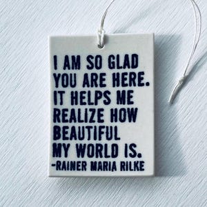 May include: A white ceramic tile with a dark blue quote from Rainer Maria Rilke: "I AM SO GLAD YOU ARE HERE. IT HELPS ME REALIZE HOW BEAUTIFUL MY WORLD IS." The tile has a small hole at the top with a white string attached.