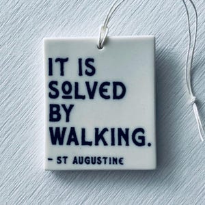 Pode incluir: Uma telha de cerâmica branca com o texto "IT IS SOLVED BY WALKING. - ST AUGUSTINE" em letras azul escuro. Uma corda branca está presa no topo para pendurar. A telha é retangular com cantos arredondados.