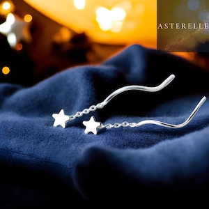 Op de afbeelding: Zilveren oorbellen met een sterontwerp. De oorbellen hebben een delicate ketting met twee sterbedels en een gebogen, golvende staaf. De oorbellen worden gepresenteerd op een donkerblauwe stof, met de merknaam "ASTERELLE" zichtbaar.