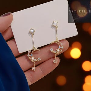Puede incluir: Pendientes dorados con diseño de luna creciente y estrella. Los pendientes tienen cristales transparentes y azules, y una pequeña perla. Los pendientes se sostienen contra una tarjeta blanca con la marca "ASTERELLE" visible.