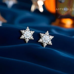 Può includere: Un paio di orecchini a forma di fiocco di neve. Ogni orecchino presenta un design a fiocco di neve argentato e dettagliato con un centro azzurro. Gli orecchini sono esposti su un tessuto blu scuro strutturato.
