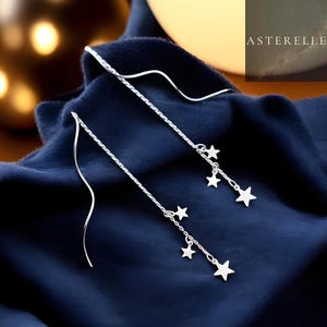May include: Silver threader earrings featuring dangling star charms. The earrings have a delicate, flowing design, with multiple stars of varying sizes suspended from the chain. The background is a deep blue fabric.