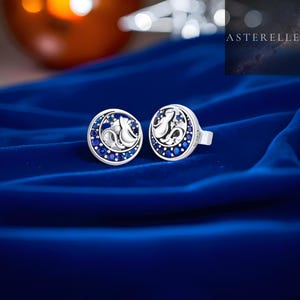May include: A pair of silver stud earrings. Each earring has a crescent moon design with a cat silhouette, surrounded by small, round, blue gemstones. The earrings are presented on a blue velvet surface.