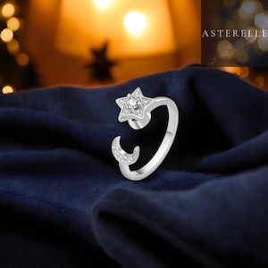 Puede incluir: Un anillo plateado con un diseño de estrella y media luna. La estrella y la luna están adornadas con pequeñas piedras brillantes. El anillo está sobre un fondo azul oscuro, con el nombre de la marca "ASTERELLE" visible en la esquina superior derecha.