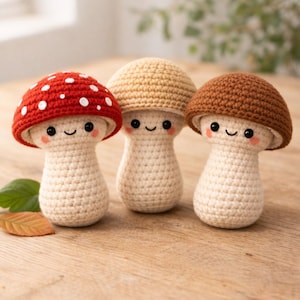 May include: Three adorable crochet mushroom figures. One has a red cap with white spots, another a tan cap, and the third a brown cap. All have cream-colored stems, smiling faces, and rosy cheeks. They are displayed on a wooden surface.