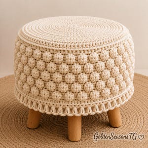 May include: A round cream-coloured crochet stool featuring a textured bobble stitch pattern on the sides and a flat top. The base is supported by three light-coloured wooden legs. The text GoldenSeasonsTG with a heart symbol is visible at the bottom.
