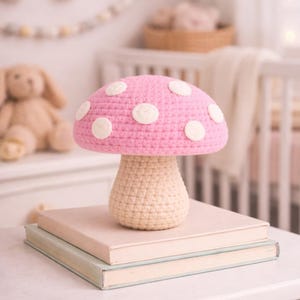 May include: A handmade crochet mushroom lamp with a pink cap and white polka dots. The mushroom sits atop a stack of books. The lamp has a beige stem and is placed on a white surface. The background includes a rainbow and a crib.