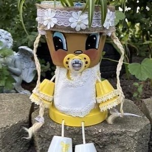 May include: A terracotta pot planter decorated to resemble a baby with a painted face, a yellow pacifier, and a white lace bib. The planter features braided yarn hair, yellow sleeves with lace trim, and white pot feet, holding a green spider plant.