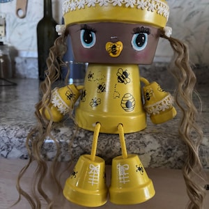 Puede incluir: Muñeca de maceta pintada a mano en amarillo y marrón. La muñeca presenta un sombrero amarillo con ribete de encaje blanco, pelo rubio largo y rasgos faciales pintados. El cuerpo está decorado con abejas y un diseño de colmena.